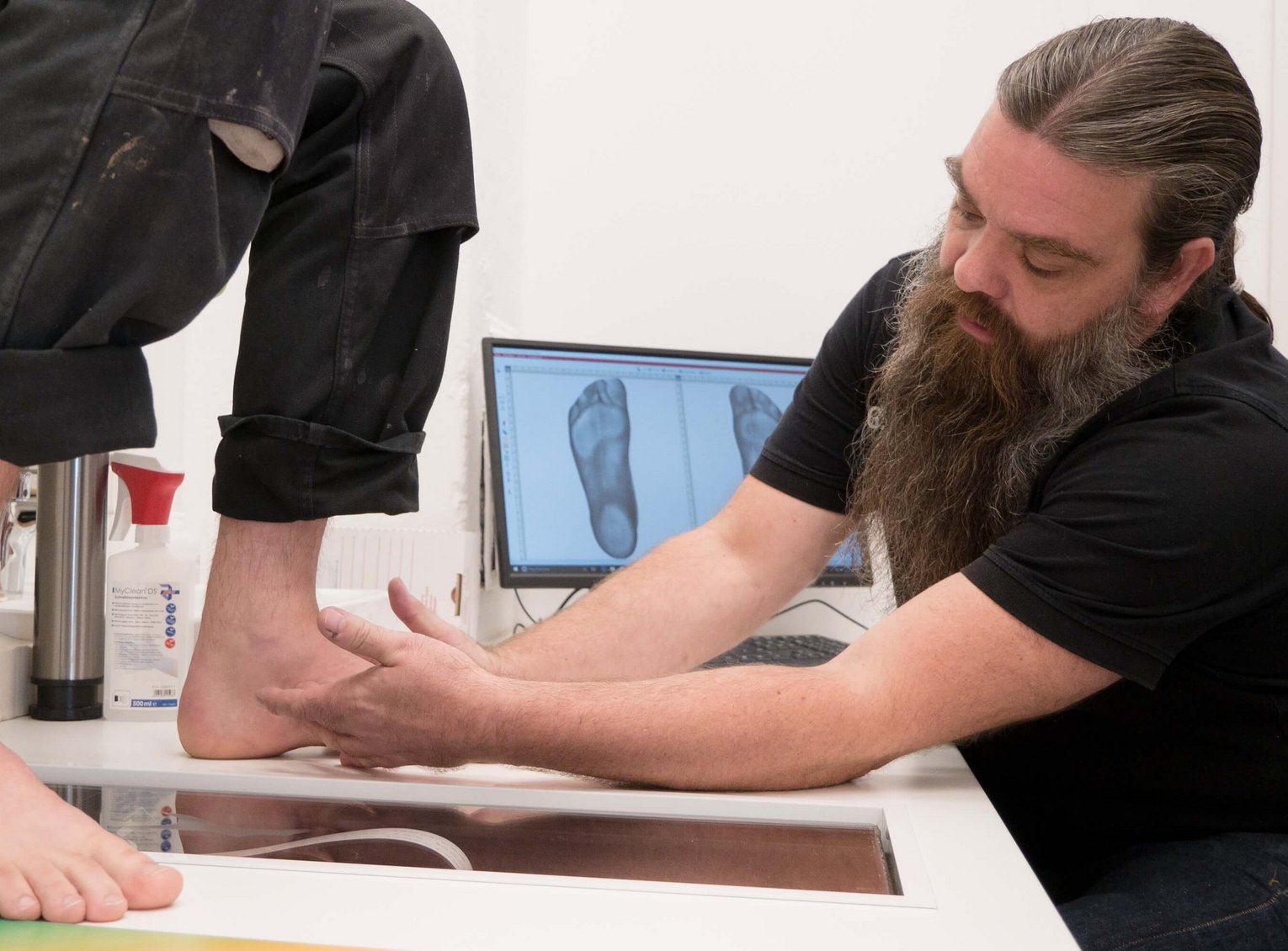 Orthopädietechniker prüft die Fußstellung eines Patienten über einem Scan-Tisch – Bildschirme im Hintergrund zeigen Fußscans